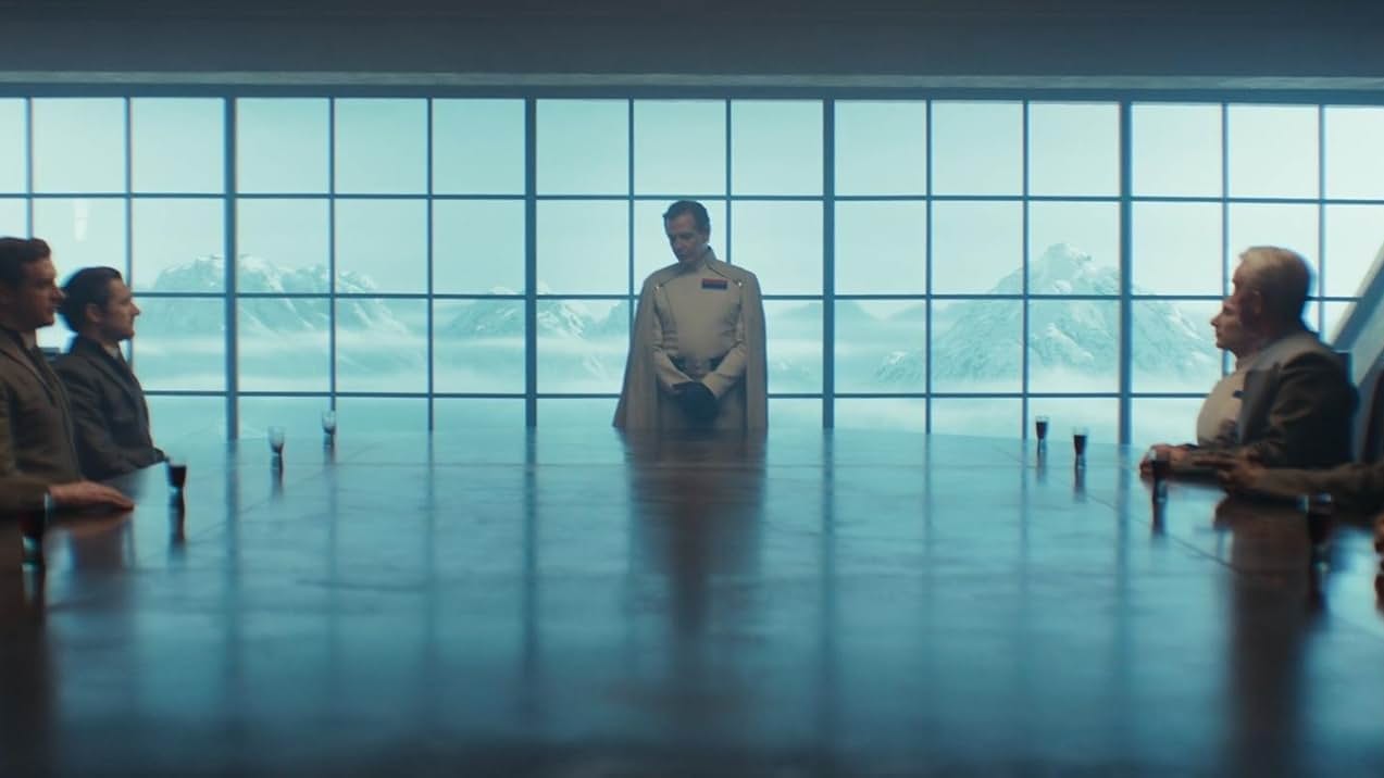 Still from the Star Wars TV show, Andor. This is the "Maltheen Divide meeting." A man in an official white uniform is talking to a select group of other officers.