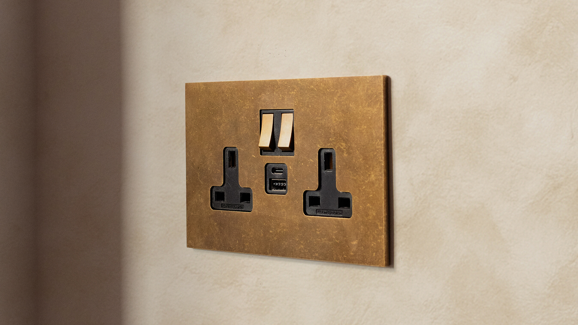 brushed matt brass twin socket on wall with peach mottled effect wallpaper behind