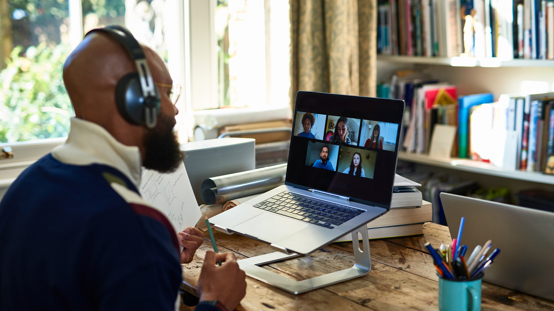 A man in headphones sits in front of the desk as he's participating in a video call on his laptop