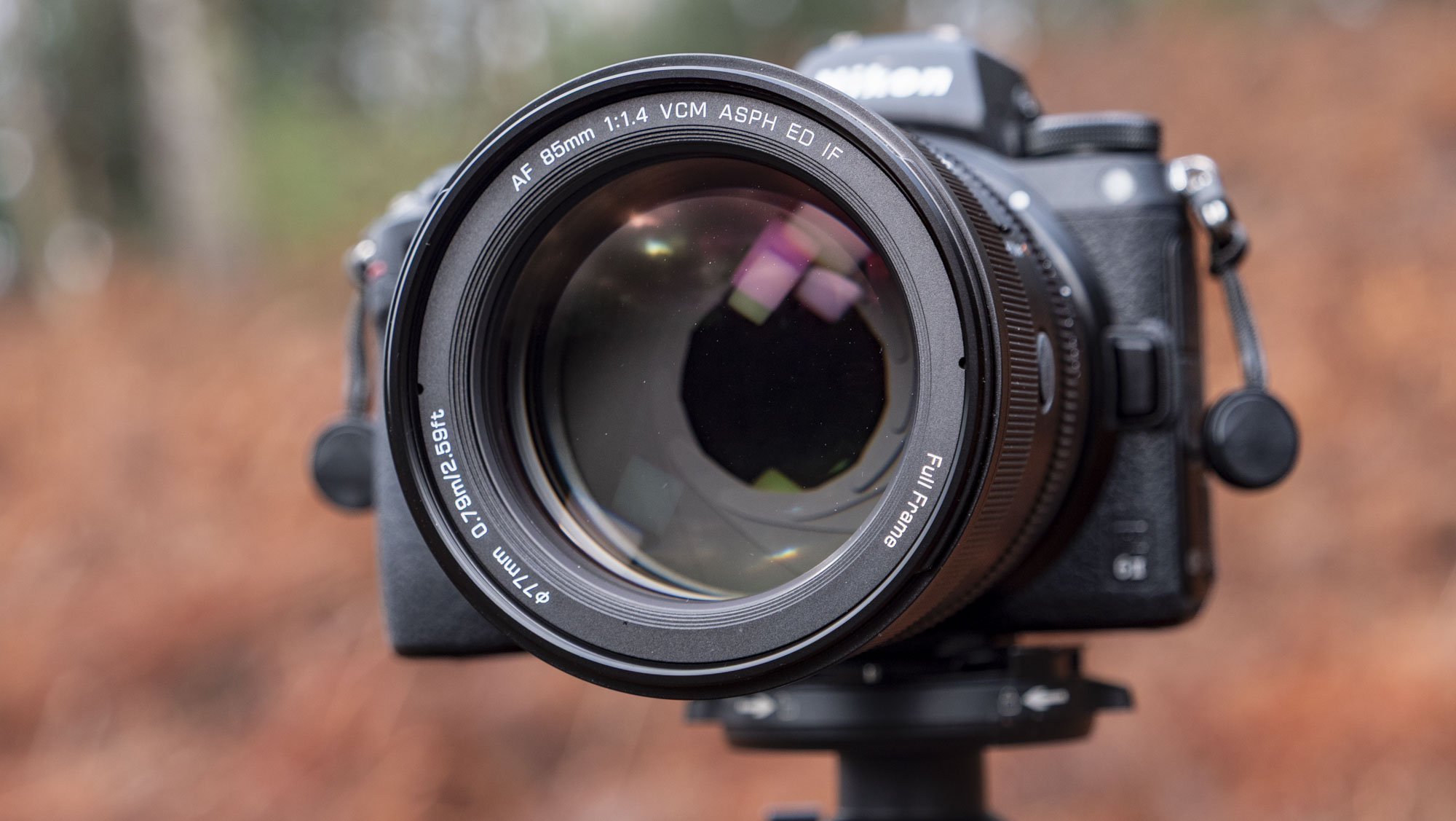 Close up of the Viltrox AF 85mm F1.4 Pro lens attached to a Nikon camera, mounted to a tripod outdoors and with bronze-colored ferns in the background, emphasis on the lens aperture blades