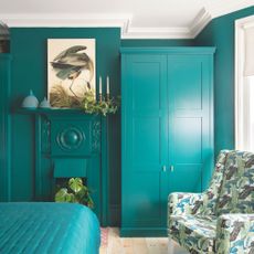 Bright teal bedroom with teal walls, a matching teal freestanding wardrobe and teal bedding, with a leaf-print accent chair beside it