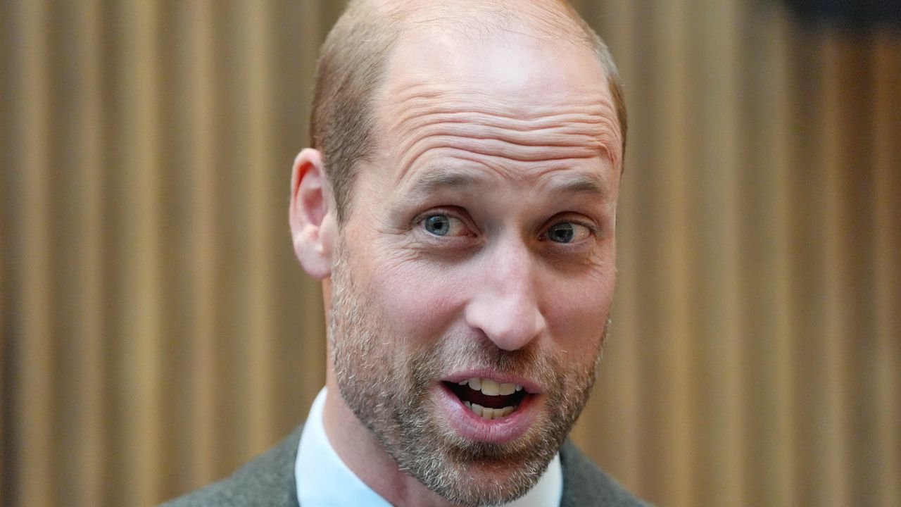 Prince William wearing a white shirt and gray blazer looking surprised while standing against a tan wall 