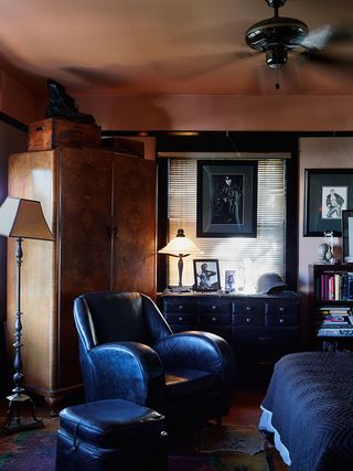 Bedroom, corner of bed with dark grey cover, black leather armchair and foot stool, wooden floor with rug, metal base standing lamp and neutral shade, dark wood wardrobe, black chest of drawers, lit lamp, photograph in a black frame, helmet, black frame window with white blinds, book case with books, framed art on walls
