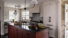 white farmhouse style kitchen with a blue patterned tile backsplash, red kitchen island with dark countertops, breakfast nook in the corner beneath the windows and white and black vintage style lighting