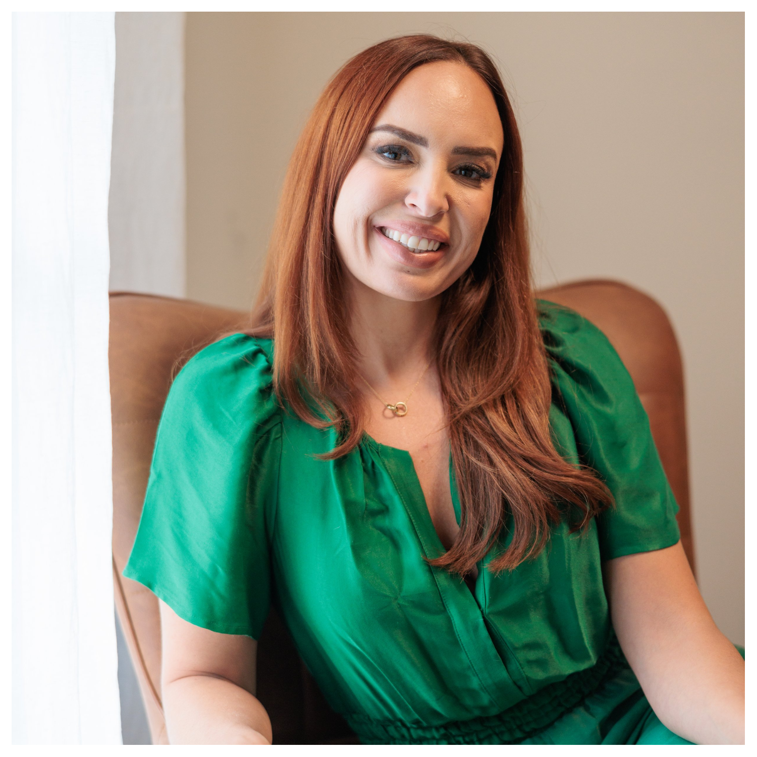 A woman with long red hair in an emerald green top sitting in a tan chair