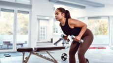 woman in a white gym space performing a standing dumbbell row with two small silver dumbbells