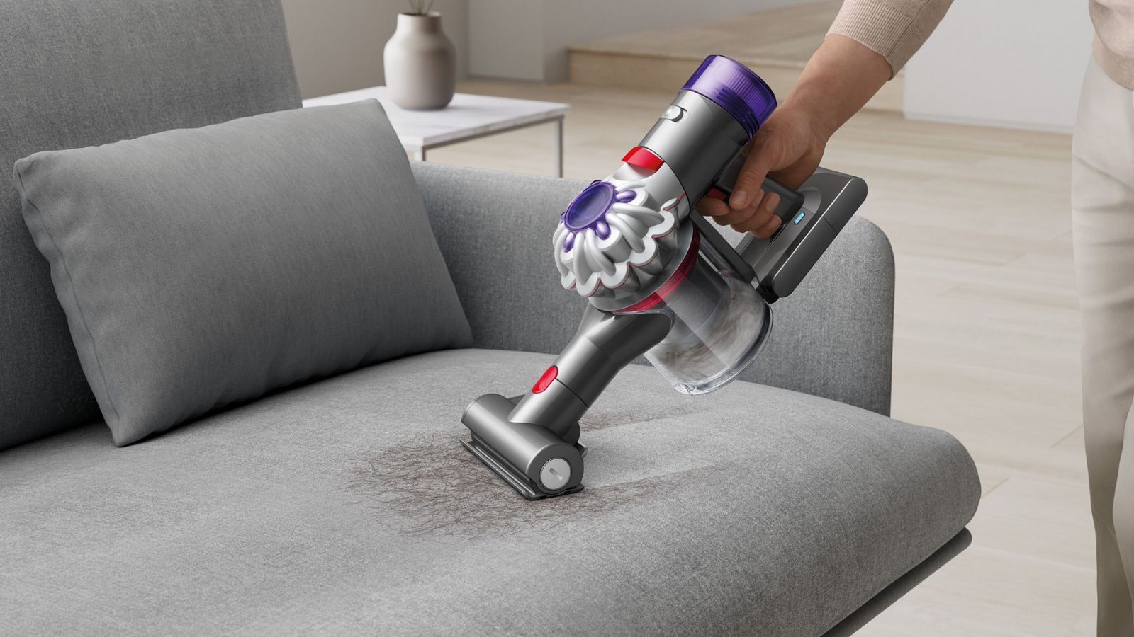 A hand using the Dyson Car+Boat handheld vacuum on a brown stain on a grey sofa, with wooden flooring beneath and a white table to the left of it, with a greige vase.