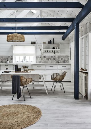 Gray plank aminate flooring in a light kitchen with wicker accents, blue painted beams and white wooden dining table.