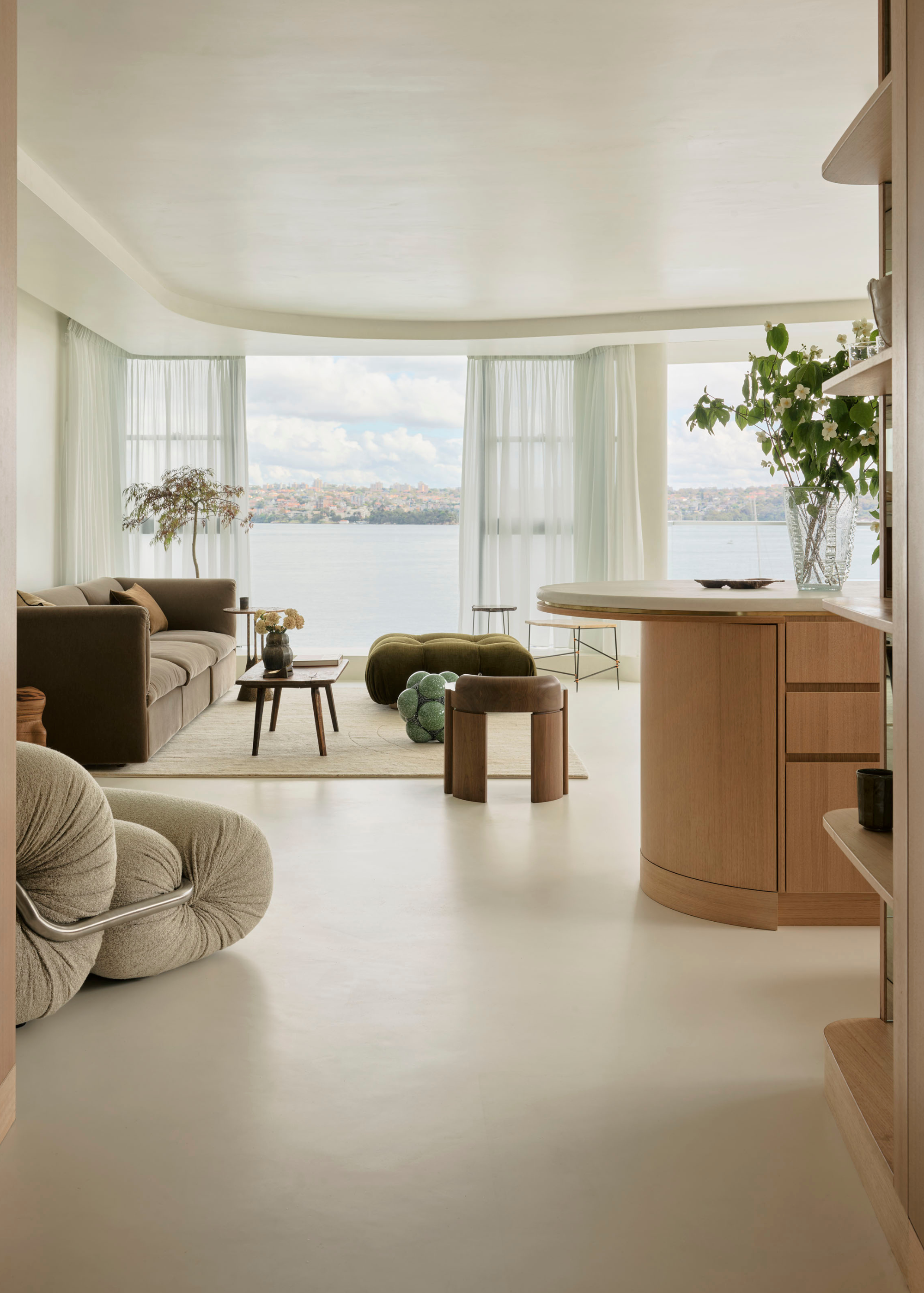 A living room with a cream sofa chair, a green pouffe, a deep muted couch, large windows, and a kitchen island