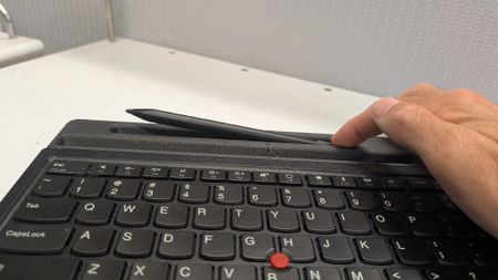 A hand places a stylus onto a keyboard's built-in holder. The keyboard is black, with a distinct red pointer, and rests on a white desk.