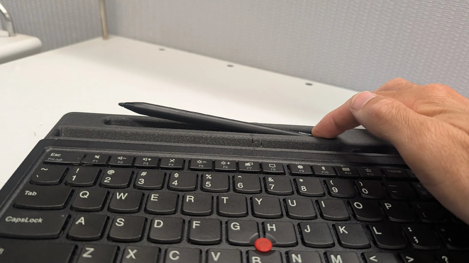 A hand places a stylus onto a keyboard's built-in holder. The keyboard is black, with a distinct red pointer, and rests on a white desk.