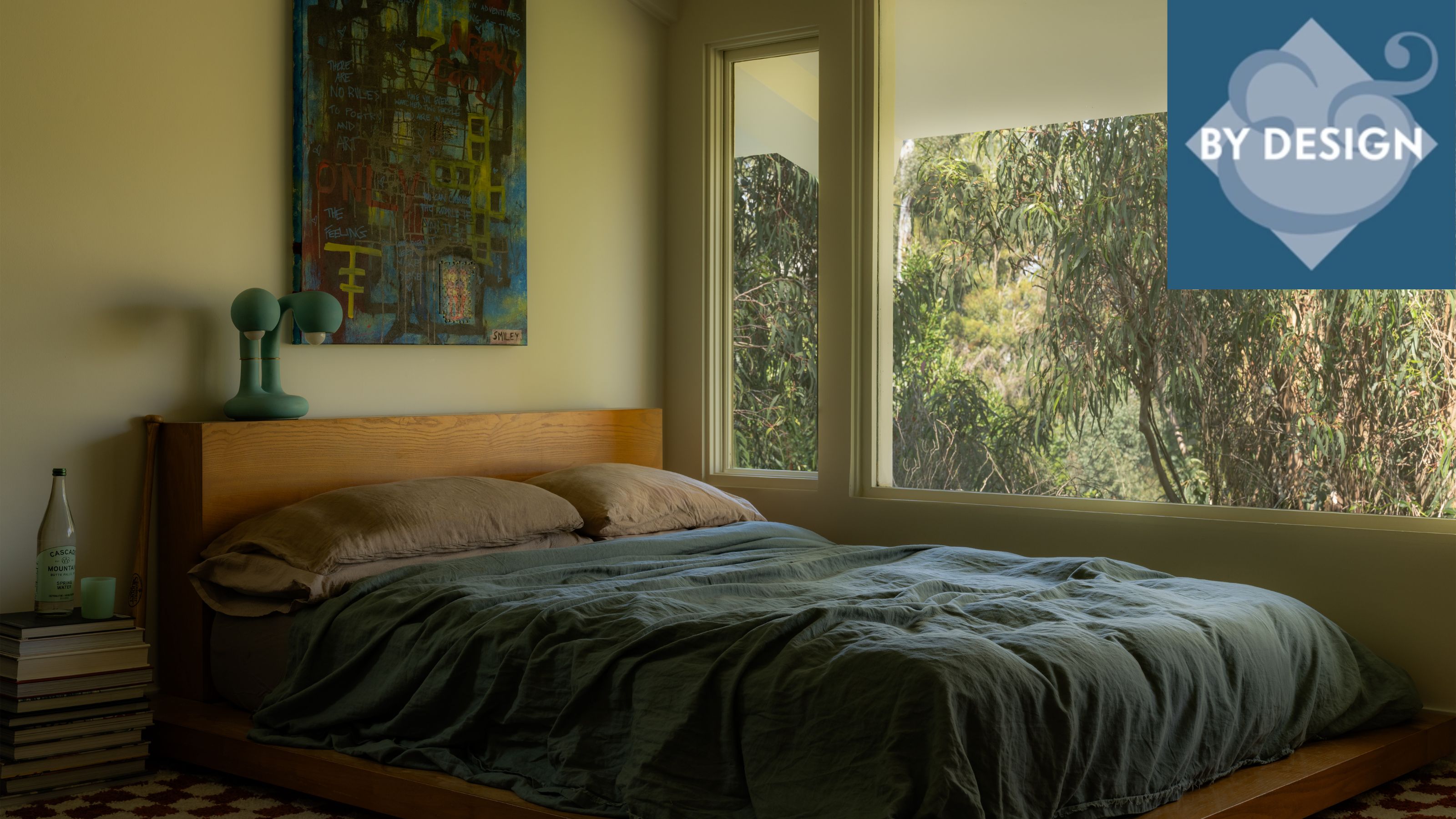 Wooden bed pushed up against a large window with blue bedding and brown pillowcases. Green walls, abstract artwork above bed, pile of books next to bed
