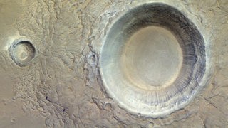 A high aerial view above a rocky, desolate landscape, with a deep, enormous crater scooped out from nearly half the image on the right. There is also a much smaller crater on the left.