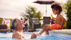 A cute grandson sits on the side of a swimming pool, holding a popsicle. He is playfully splashing his smiling grandfather.