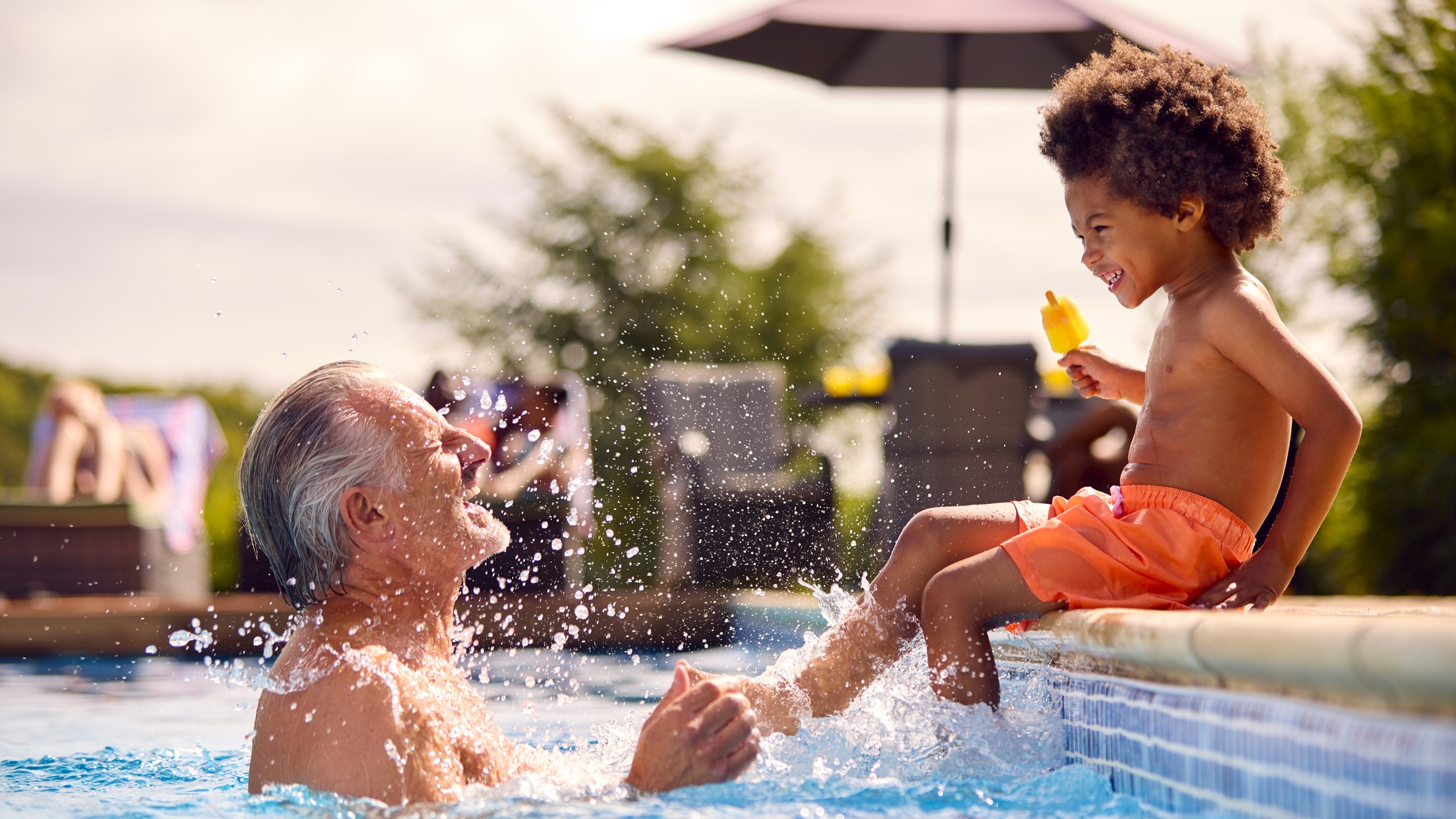 A cute grandson sits on the side of a swimming pool, holding a popsicle. He is playfully splashing his smiling grandfather.