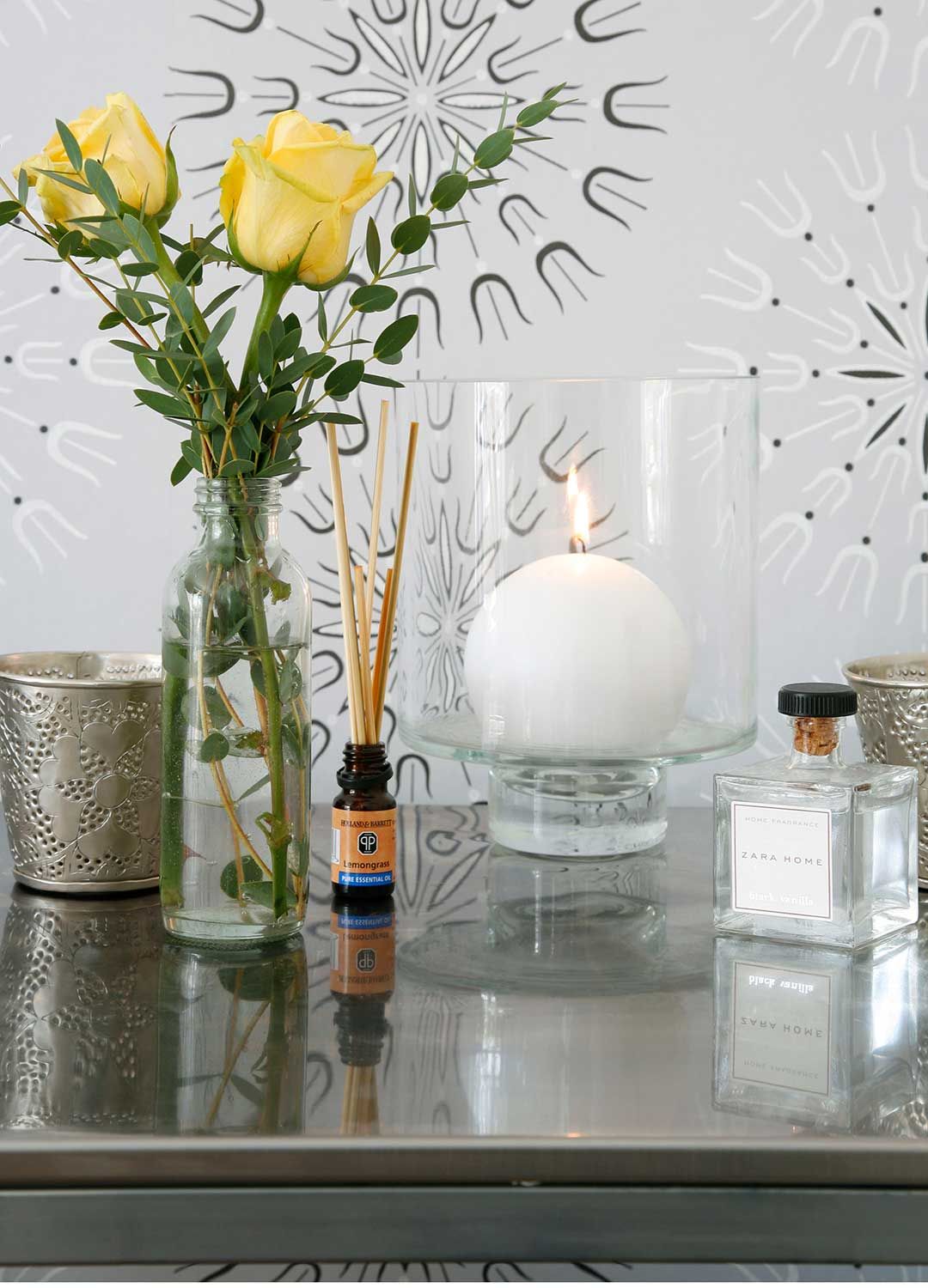 homes fragrances on a glass sideboard with yellow roses in a vase in front of a sunburst wallpaper design