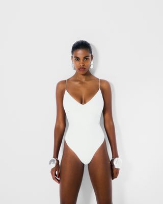 A Reformation model wears a white Reformation swimsuit in front of a plain backdrop