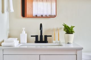 A bathroom sink with black faucets, on top of it are two bamboo toothbrushes and a white pot with a plant on the right, and two white folded towels and a white soap dispenser on the left
