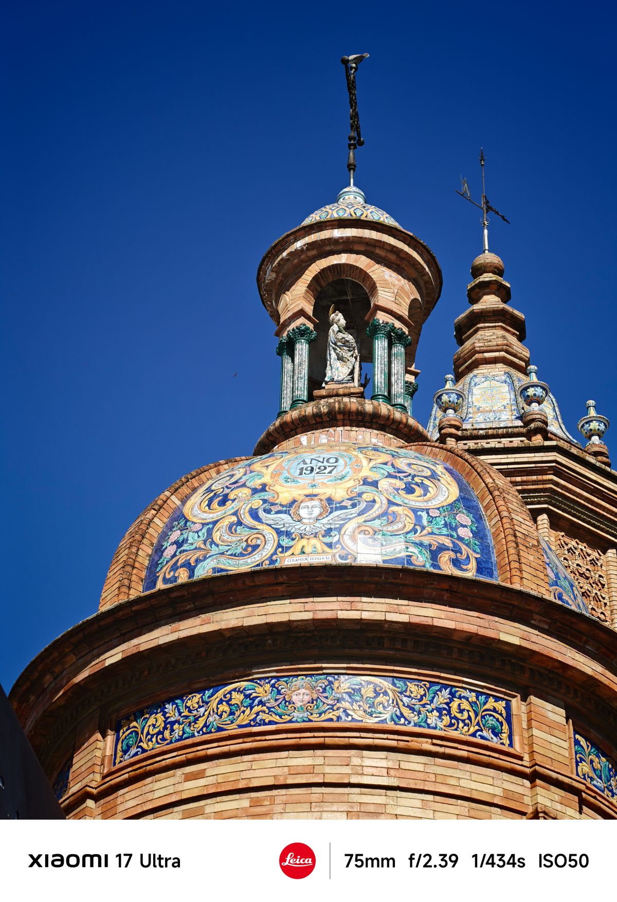 Ornate tiled dome with intricate blue and gold patterns and a small statue at the top against a vivid blue sky.