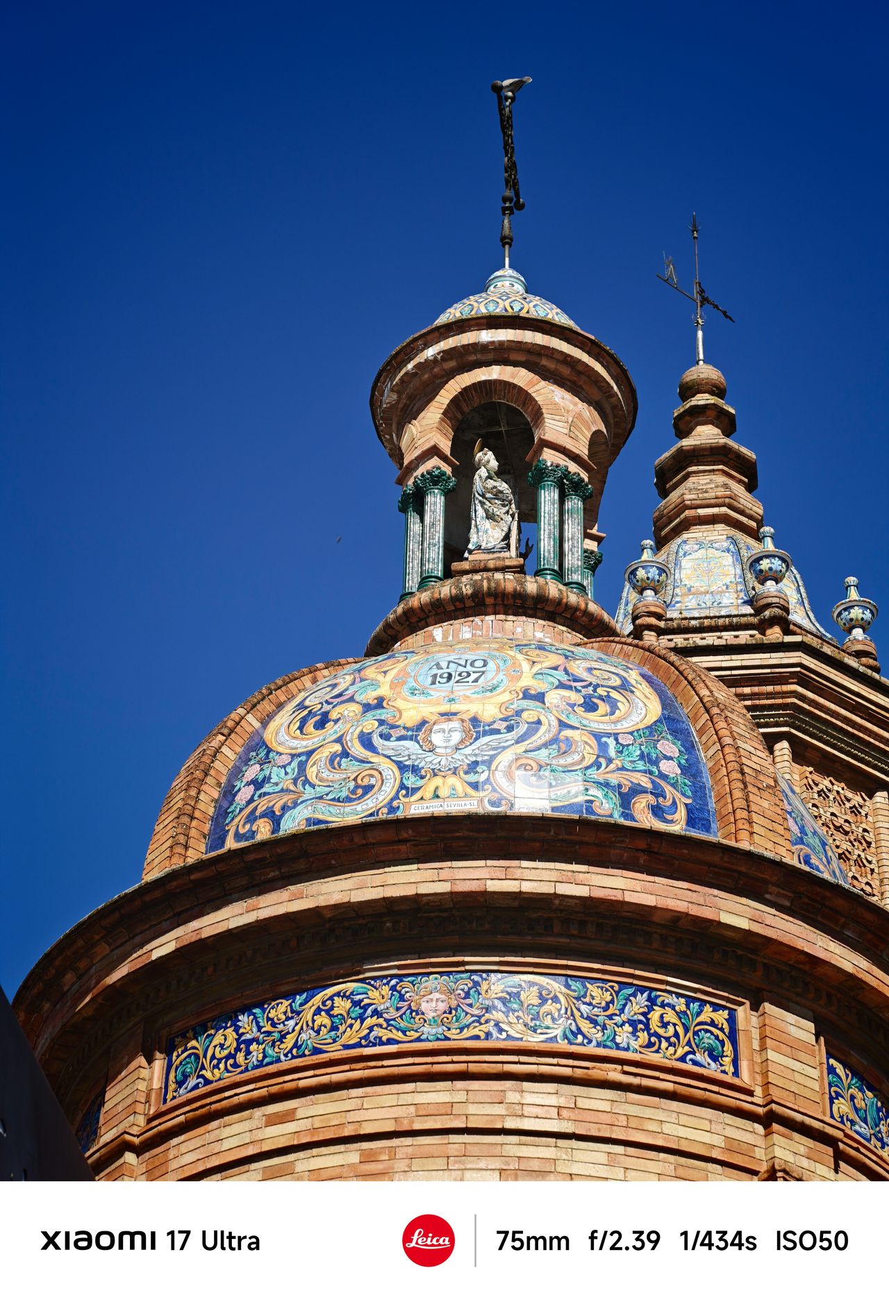 Ornate tiled dome with intricate blue and gold patterns and a small statue at the top against a vivid blue sky.