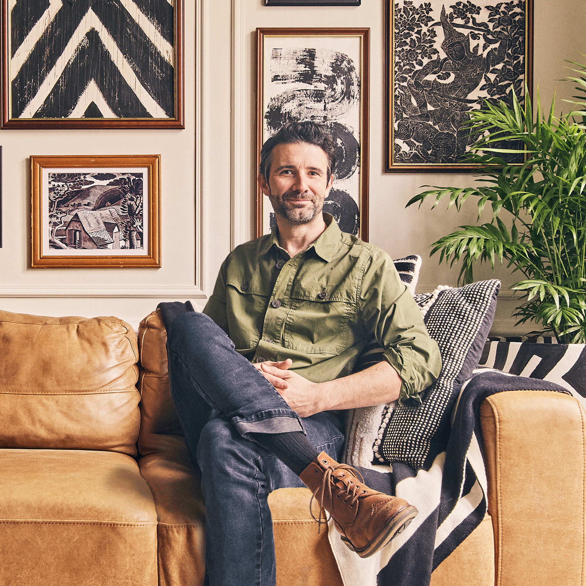 Interior designer John Coper sitting on a brown leather sofa with a gallery wall backdrop wearing a green shirt and dark denim jeans