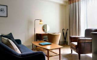 A room in the Redchurch Townhouse hotel. A deep blue couch and a brown leather armchair have a wooden table in between them, while the antique-looking wooden des sits against a wall with a light brown chair. The window lets in a lot of light.