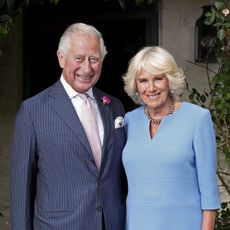 King Charles in a striped blue suit standing next to Queen Camilla, in a blue dress, and smiling 