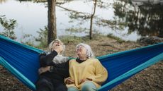 Two older women share a hammock and laugh on the side of a lake.