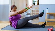 A woman performs seated leg lifts at home on an exercise mat. She is sitting on the mat, with her left leg resting on the mat and her right leg pointing up. Her arms are raised and a resistance band is looped around her wrists.