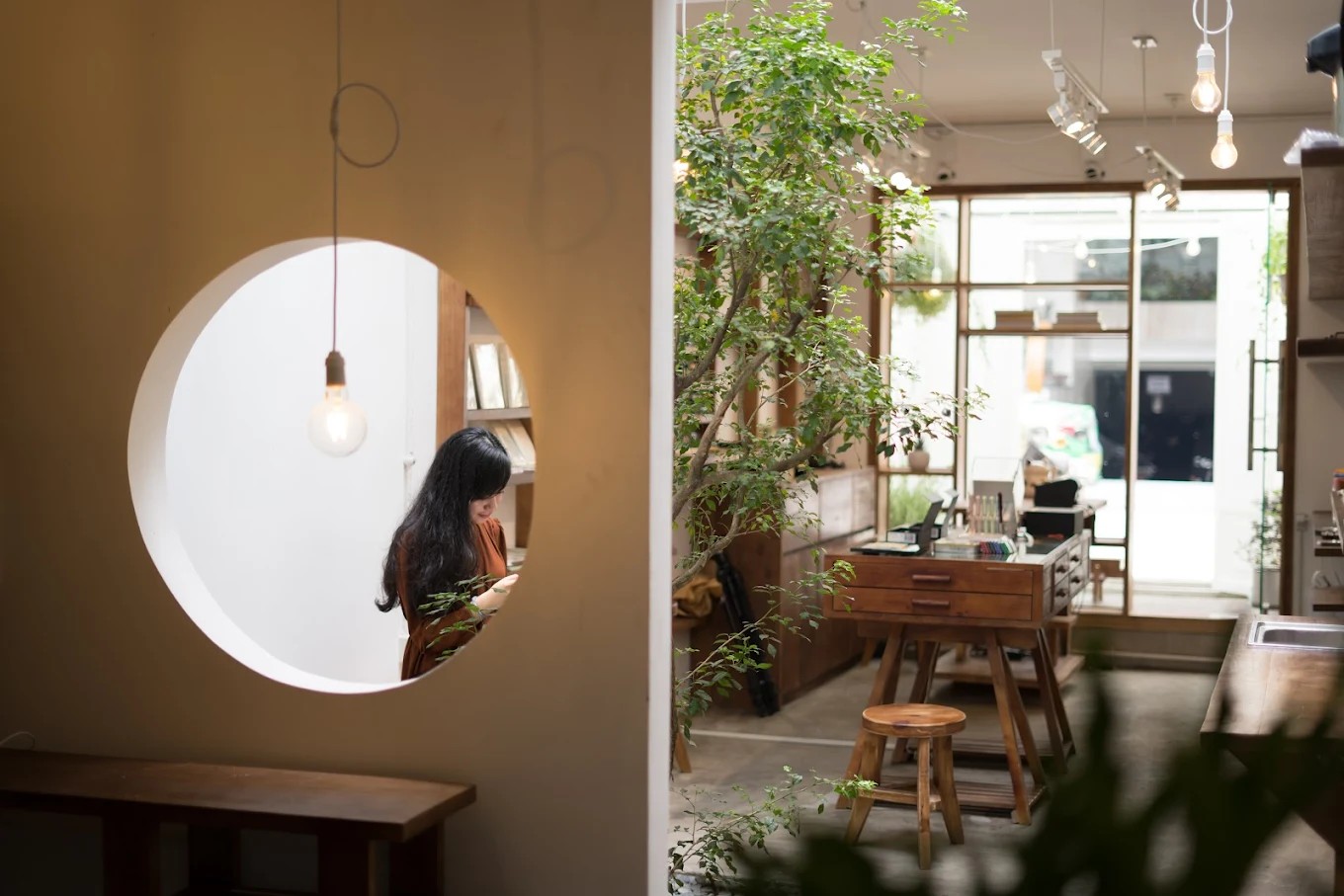 A wood-sculpted, minimalist bookstore with plants and vintage furniture features a see-through porthole window behind which a woman reads.