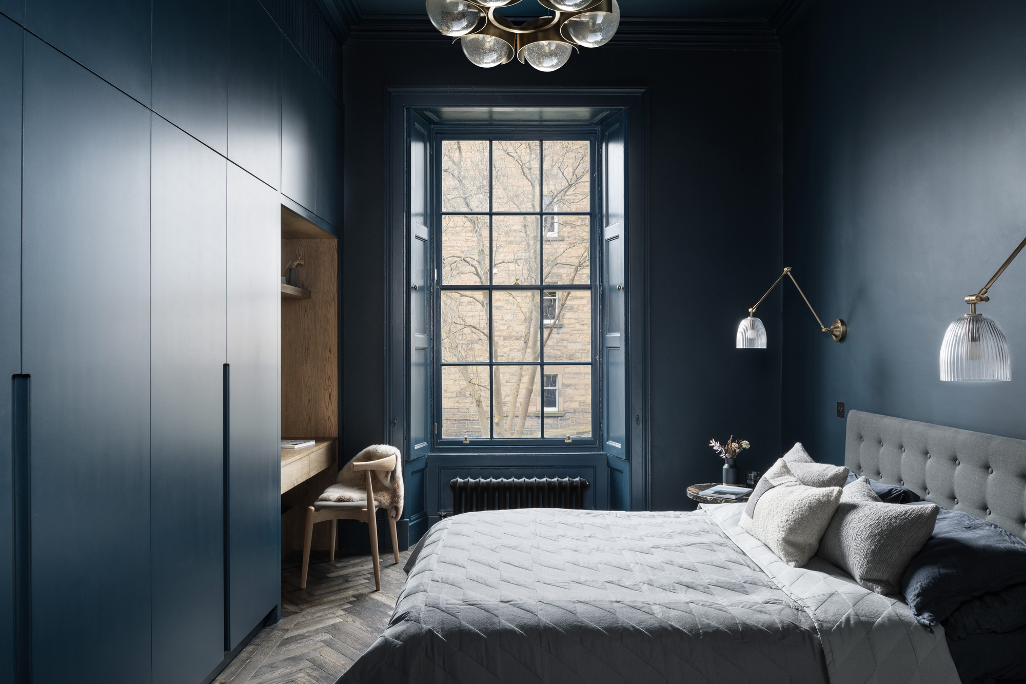 dark navy blue bedroom with large window, radiator under it, built-in blue cabinetry with a timber desk built-in and timber chair, color drenched walls, two wall sconces on either side of the bed, gray headboard, blue and gray sheets