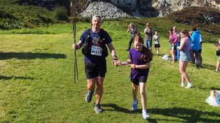 A man runs through a field at the end of the race, holding hands with a young girl who is pulling him forwards to finish.