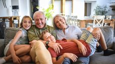 A pair of happy grandparents lounge on the couch with their grandkids snuggled close. They appear wealthy and relaxed.