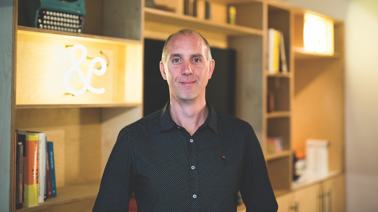 A photo of web typography expert Richard Rutter standing in front of pine shelves. An ampersand made out of flourescent tubing is clearly visible behind him.