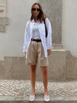 a photo of a woman wearing a white button-down shirt over a tank top with pleated shorts and mesh ballet flats