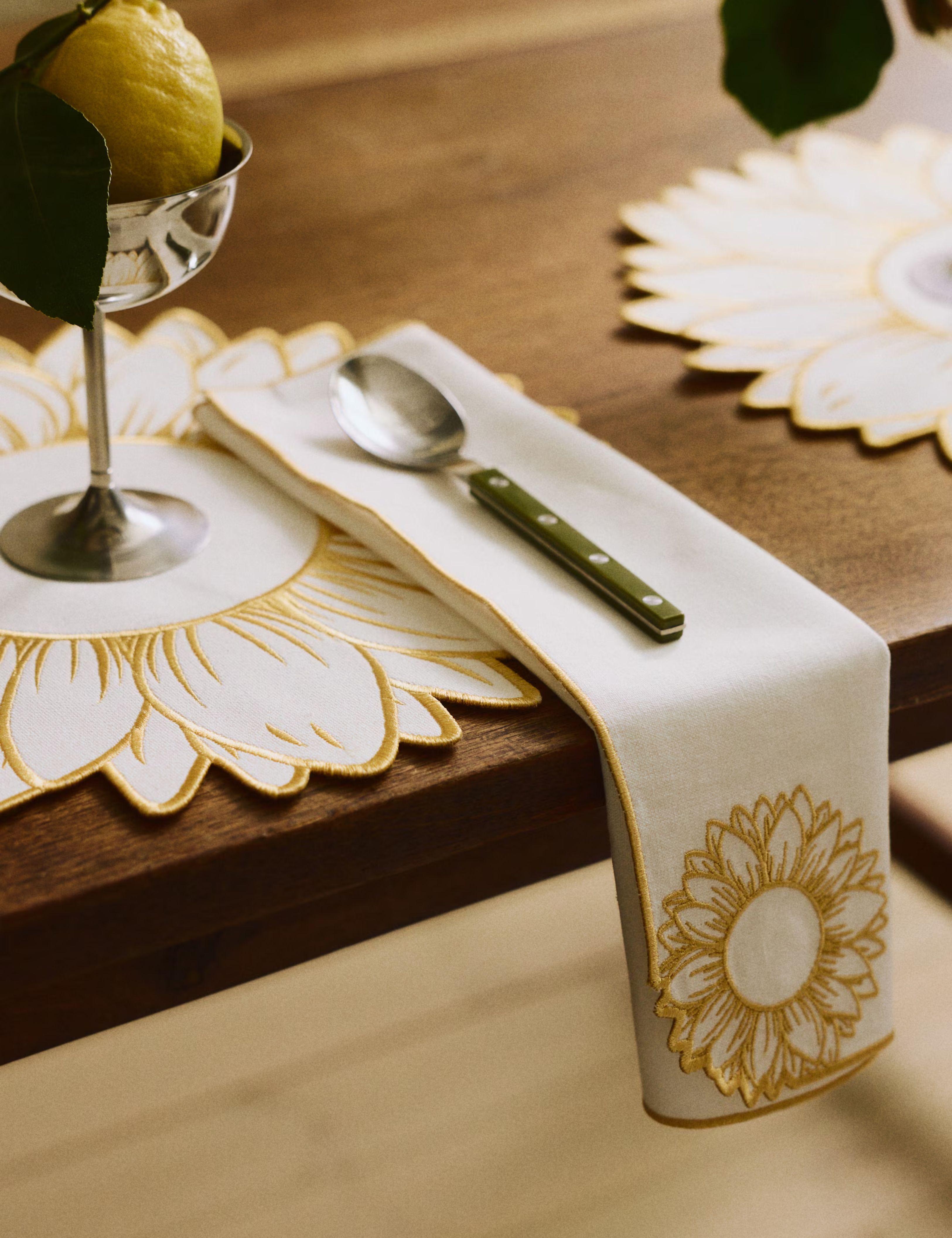 White sunflower-embroidered table linens styled with chrome cutlery and stemware atop of a wooden table
