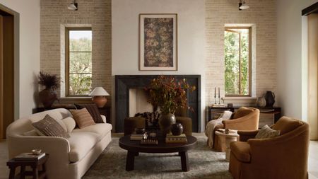 Warm neutral living room with cream curved sofa, a dark wood coffee table, two velvet brown armchairs, a large fireplace and a floral print above.