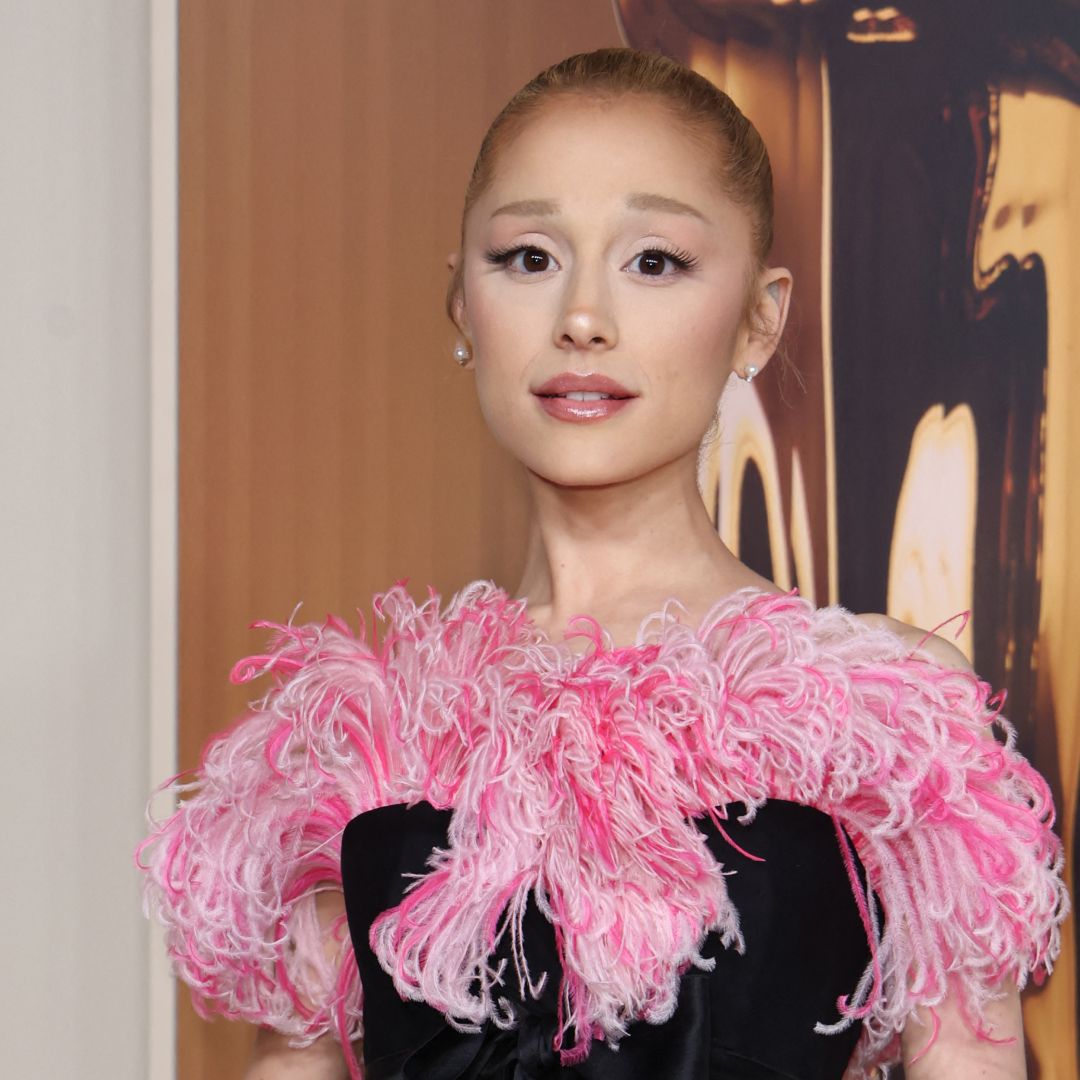 Ariana Grande wearing a slicked back ponytail as well as a black dress that has pink and white feathers at the neck line. 