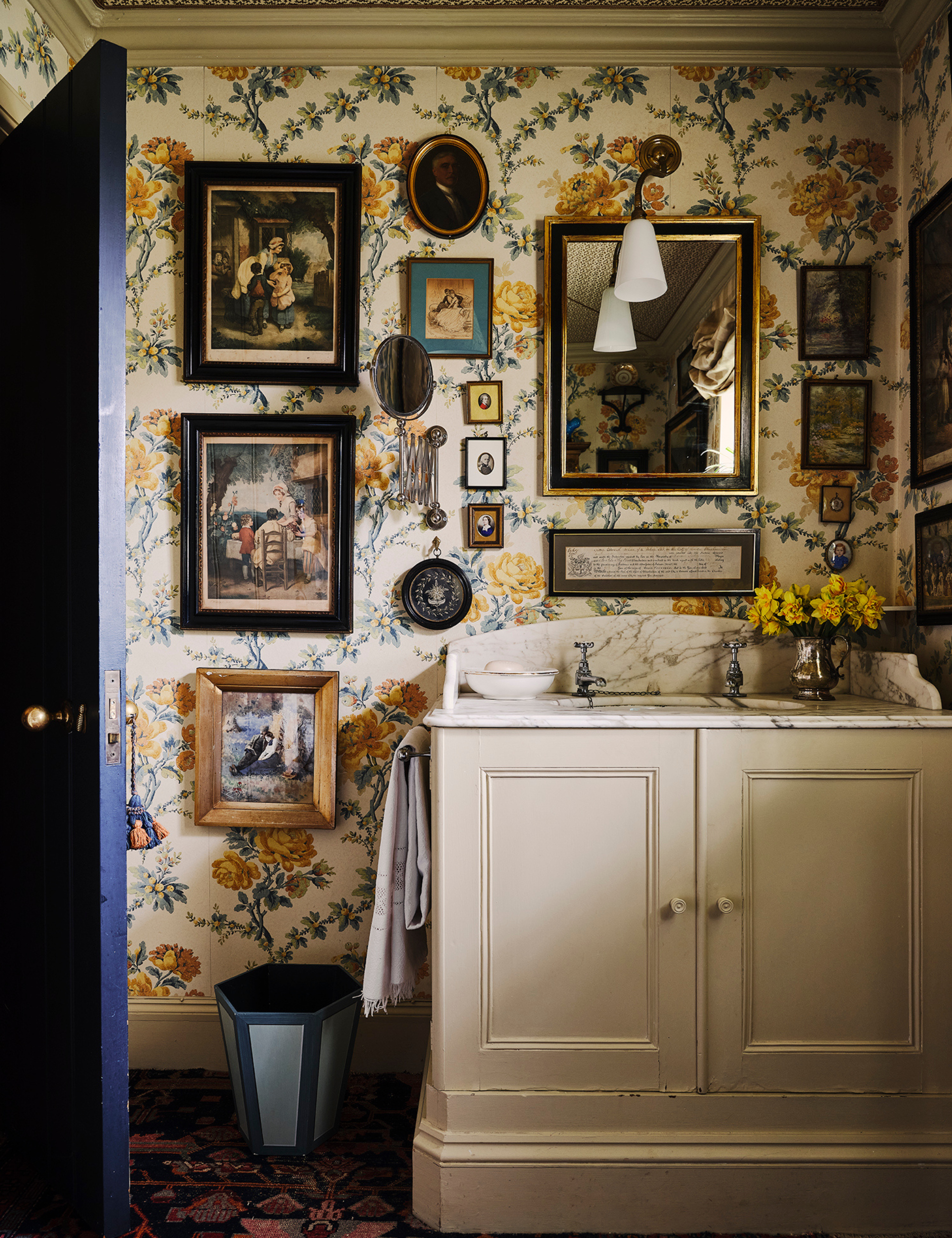 Collected vintage-style bathroom featuring ample wallpaper, a tonal green wastebasket, and a vase full of yellow flowers