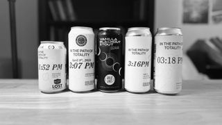 Five cans stand on a wooden table against a blurred, non descript homey backdrop. The middle can is black with a diagonal solar eclipse progression illustrated. the other cans are white, with times on them. the leftmost can is shorter than the others. 
