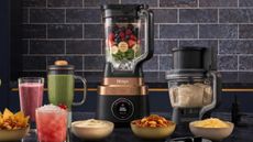 The ninja CrushBOSS blender with a food processor and a smoothie cup attachment either side of it, against black wall times. 
