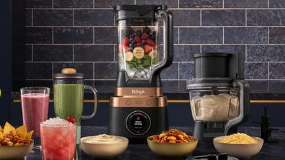 The ninja CrushBOSS blender with a food processor and a smoothie cup attachment either side of it, against black wall times.