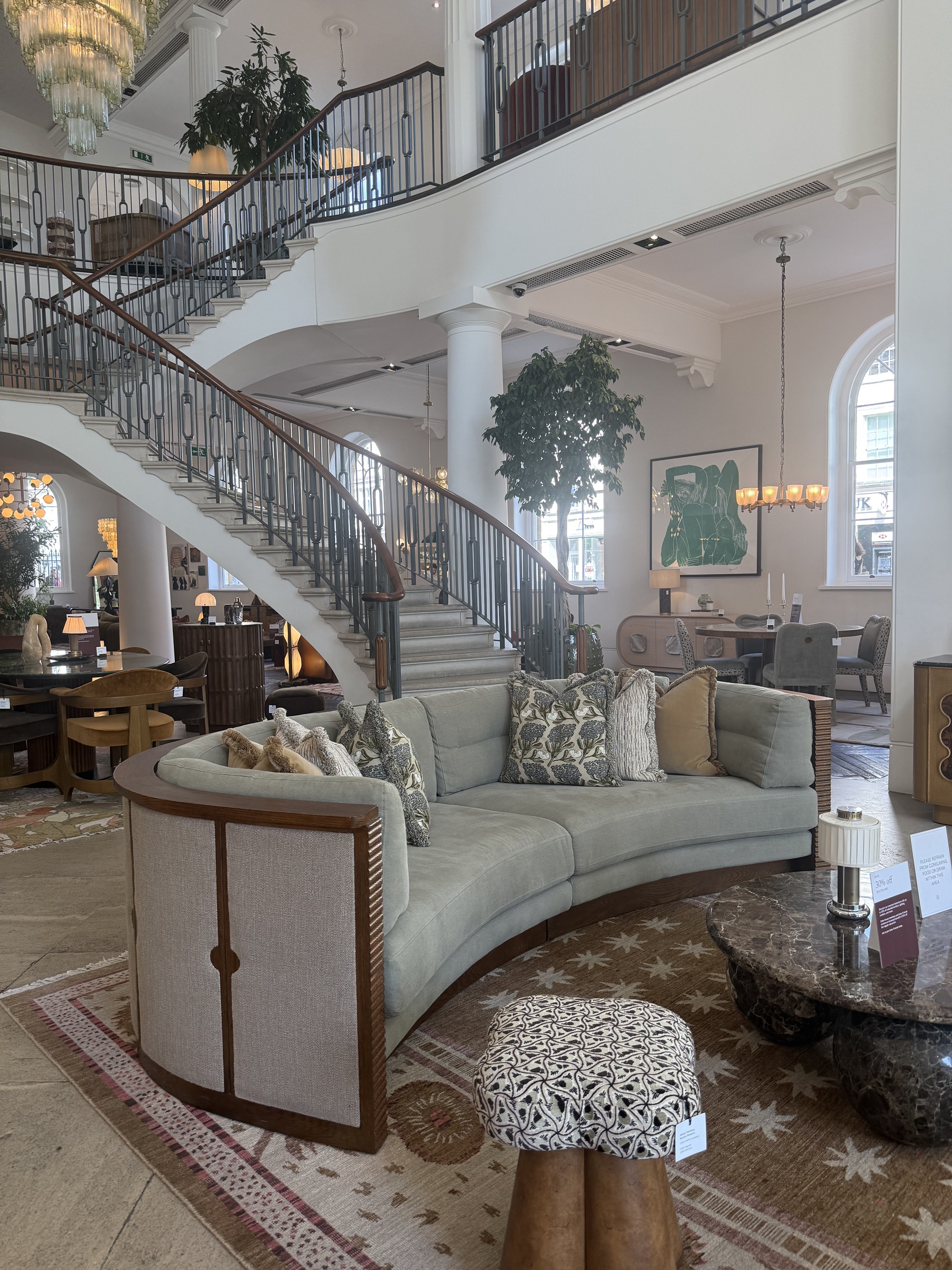 soho home sofa in showroom in front of grand staircase, with fabric and timber backing, curved shape styled with cushions on patterned rug with ottoman and stone coffee table