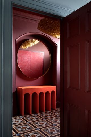 Hallway with walls in rich burgundy paint, tiled burgundy floor and red sculptural console table below large round mirror in arched alcove