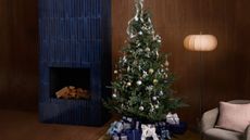 christmas tree with lots of presents beneath next to a blue tiled fireplace, timber panelled wall, armchair and floor lamp