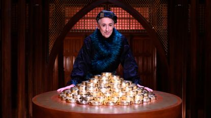 Host Alan Cumming leans on a round table with a pile of large gold coins, while standing in a chamber, in &#039;The Traitors&#039; season 3.