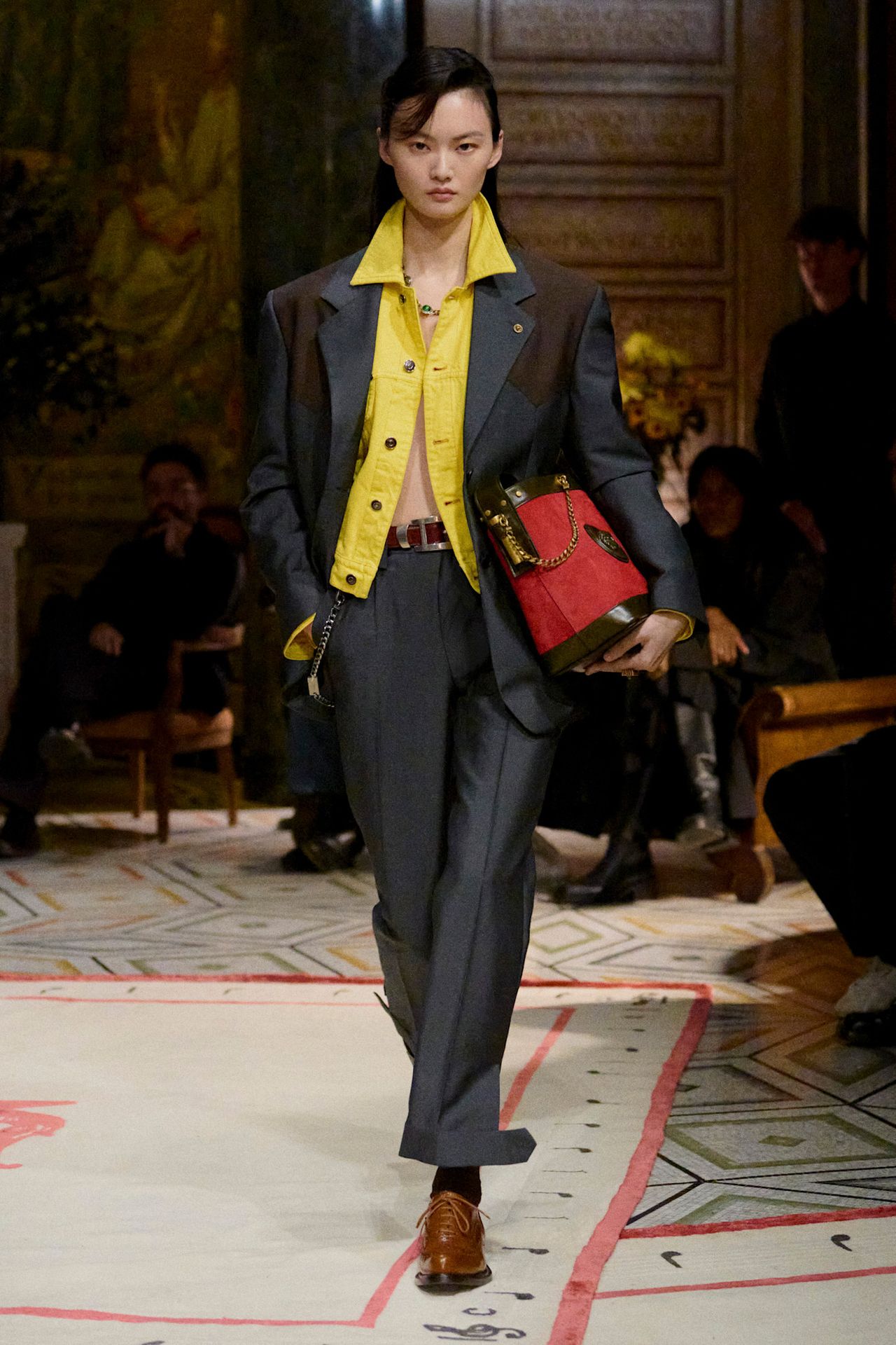 Layering staples for spring 2026 is shown in a photo from Versace's runway show, with a model walking while wearing an oversized gray and brown two-tone blazer styled over a bright yellow denim jacket worn with matching gray trousers, a brown belt, and brown oxford shoes, black socks, and a red suede bucket bag
