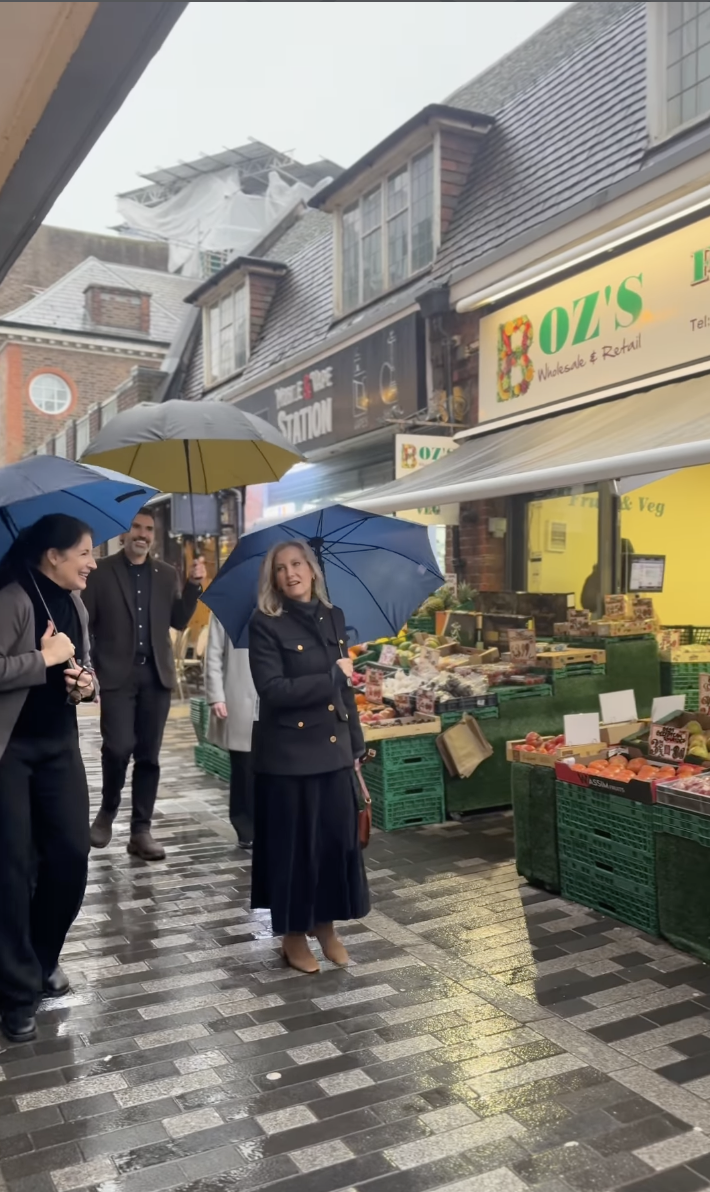 Duchess Sophie standing outside a fruit stand with a man and woman, holding an umbrella