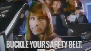 Iron Maiden singer Bruce Dickinson sat in a convertible and looking at the camera, with text reading "Buckle your safety belt."