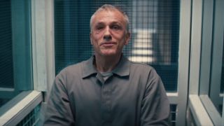 A scarred Christoph Waltz smiles while sitting in his jail cell in No Time To Die.
